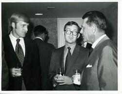 ["Black and white photograph print of three unidentified men talking with one another."]