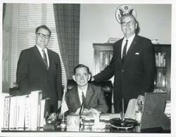 ["Black and white photograph print of Carl Albert seated at his desk. Two unidentified men are standing behind him."]