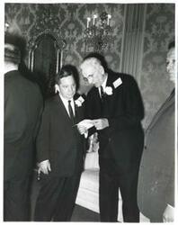 ["Black and white photograph print of Carl Albert and John W. McCormack looking at a document. Other people are around them."]