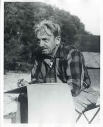 ["Black and white photograph print of Thomas Hart Benton sitting outside. He is writing something or making a sketch. October 12, 1973"]