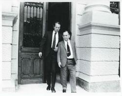 ["Black and white photograph print of Carl Albert walking with an unidentified man outside a building during the House delegation trip. August 1975"]