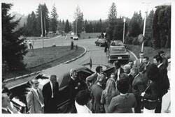 ["Black and white photograph print of several people standing outside waiting to be escorted away in cars. House delegation trip. August 1975"]