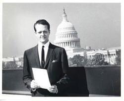 ["Black and white photograph print of an unidentified man standing outside. The Capitol is in the background."]