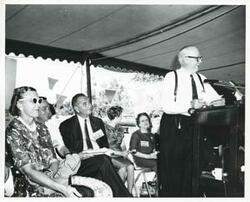 ["Black and white photograph print of Robert S. Kerr speaking at a podium. Seated behind him is Mrs. Kerr, Carl Albert, and Lyndon B. Johnson."]