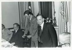 ["Black and white photograph print of Carl Albert standing in a room with several other people."]
