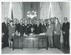 ["Black and white photograph print of Carl Albert posing with several people behind his desk."]