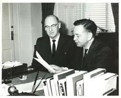 ["Black and white photograph print of Carl Albert speaking with an unidentified man. They are looking at documents."]