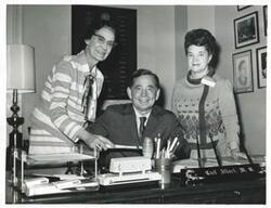 ["Black and white photograph print of Carl Albert seated at his desk. Two unidentified women are standing behind him."]
