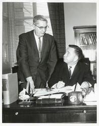 ["Black and white photograph print of Carl Albert seated at his desk. An unidentified man is standing behind him."]