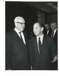 ["Black and white photograph print of Carl Albert speaking with an unidentified man. Wright Patman is in the background."]