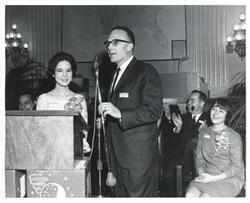 ["Black and white photograph print of Mary Frances Albert standing at a podium. There is a man standing beside her."]