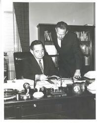 ["Black and white photograph print of Carl Albert seated at his desk with a man standing behind him. They are looking at a book."]
