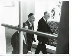["Black and white photograph print of Carl Albert, Mike Mansfield, and John W. McCormack walking together outside."]
