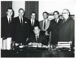 ["Black and white photograph print of Carl Albert seated at his desk. Standing behind him L to R are: Jed Johnson, Jr., A. S. Mike Monroney, Ed Edmondson, John Jarman, Fred Harris, and Tom Steed."]