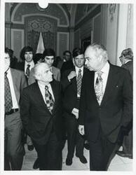 ["Black and white photograph print of Robert Strauss, Jonathan Brigham, and others walking down a corridor."]