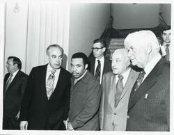 ["Black and white photograph print of Robert Strauss, Thomas P. O'Neill, and others standing in a corridor."]