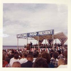 ["Black and white photograph print of John B. Connally, A. S. Mike Monroney, Lady Bird Johnson, Henry Bellmon, Carl Albert, Ed Edmondson, Lyndon B. Johnson, and others at the dedication of the Eufaula Dam and Reservoir by President Johnson. September 25, 1964. a-c color photo additions. Carl Albert at podium, Lyndon B. Johnson at podium, and crowd."]