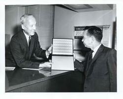 ["Black and white photograph print of Carl Albert with an unidentified man looking at the Sam Rayburn commemorative stamp."]