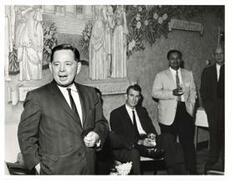 ["Black and white photograph print of Carl Albert speaking with three unidentified people in the background"]