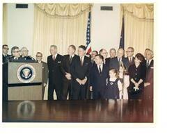 ["Color photograph print of swearing-in ceremonies for Postmaster General John A. Gronouski, who is speaking.  Also present (from left to right): Henry J. Stoffer (President of the National League of Postmasters); Chester W. Parrish (Secretary-Treasurer of the National Federation of Post Office Motor Vehicle Employees, AFL-CIO); Don E. Dunn (Executive Vice-President of the United Federation of Postal Clerks, AFL-CIO); Frederick J. O'Dwyer (President of the National Association of Postal Supervisors); Senator A. Willis Robertson of Virginia; Attorney General Katzenbach; Representative John A. Race of Wisconsin; House Majority Whip Hale Boggs (of Louisiana); Representative Clement J. Zablocki of Wisconsin; Carl Albert  (as House Majority Leader); Representative Henry S. Reuss of Wisconsin; Representative Lynn Stalbaum of Wisconsin; Senator A. S. Mike Monroney of Oklahoma; Representative Melvin R. Laird of Wisconsin; Secretary of Labor W. Willard Wirtz; Mrs. Gronouski; Secretary of the Treasury C. Douglas Dillon; Senator Frank Carlson of Kansas; and Postmaster General's daughters Julie and Stacy Gronouski (in the foreground). February 19, 1965"]