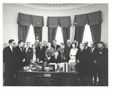 ["Black and White photograph, on stock paper, of John F. Kennedy signing S. 1922, the Housing Bill. Carl Albert and Lyndon B. Johnson are present."]