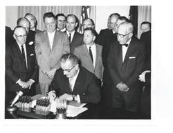 ["Black and white photograph, on stock paper, of Lyndon B. Johnson signing the cotton-wheat bill in president's office. Pictured are George D. Aiken, Carl Albert, Hale Boggs, Orville Freeman, Hubert H. Humphrey, George S. McGovern, and W.R. Poage."]