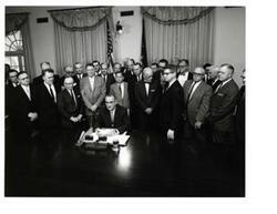 ["Black and white photograph of Lyndon B. Johnson signing the cotton-wheat bill in president's office. Pictured are George D. Aiken, Carl Albert, Hale Boggs, Orville Freeman, Hubert H. Humphrey, George S. McGovern, and W.R. Poage."]