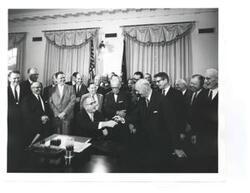 ["Black and white photograph, on stock paper, of Lyndon B. Johnson signing the cotton-wheat bill in president's office. Pictured are George D. Aiken, Carl Albert, Hale Boggs, Orville Freeman, Hubert H. Humphrey, George S. McGovern, and W.R. Poage."]
