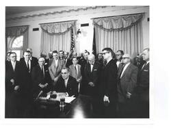 ["Black and white photograph, on stock paper, of Lyndon B. Johnson signing the cotton-wheat bill in president's office. Pictured are George D. Aiken, Carl Albert, Hale Boggs, Orville Freeman, Hubert H. Humphrey, George S. McGovern, and W.R. Poage."]
