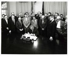 ["Black and white photograph of Lyndon B. Johnson signing the cotton-wheat bill in president's office. Pictured are George D. Aiken, Carl Albert, Hale Boggs, Orville Freeman, Hubert H. Humphrey, George S. McGovern, and W.R. Poage. Handwritten message identifying event, by unknown author, on back of photo."]