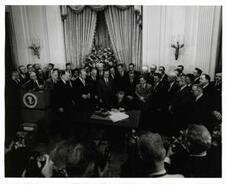["Black and white photograph of Lyndon B. Johnson signing the tax bill in the president's office. Those pictured include: Carl Albert, Everett M. Dirksen, Lady Bird Johnson, Russell B. Long, John McCormack, and Wilbur D. Mills."]