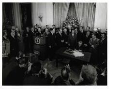 ["Black and white photograph of Lyndon B. Johnson signing the tax bill in the president's office. Those pictured include: Carl Albert, Everett M. Dirksen, Lady Bird Johnson, Russell B. Long, John McCormack, and Wilbur D. Mills. Handwritten message identifying event, by unknown author, on back of photo."]