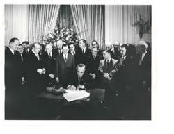 ["Black and white photograph, on stock paper, of Lyndon B. Johnson signing the tax bill in the president's office. Those pictured include: Carl Albert, Everett M. Dirksen, Lady Bird Johnson, Russell B. Long, John McCormack, and Wilbur D. Mills. Handwritten message identifying event, by unknown author, on back of photo."]