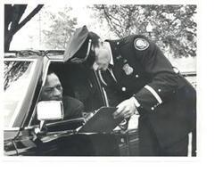 ["Black and white photograph of Carl Albert's (unknown) driver speaking to a security clerk with Carl Albert in the back of the limousine"]