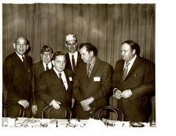 ["Black and white photograph of Carl Albert posing with four unidentified men at a dinner connected to a Disabled American Veteran state convention. Pictured center (background) is D.A.V. Representative  Paul Freeman. 1970."]