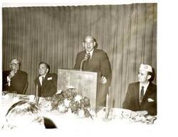 ["Black and white photograph of Carl Albert sitting at the speaker's table, next to an unidentified man speaking at the event on his right (our left), at a Disabled American Veterans state convention. On the other side of the event speaker is D.A.V. Representative  Paul Freeman. 1970."]