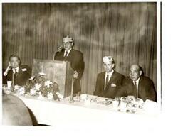 ["Black and white photograph of Carl Albert sitting at the speaker's table, next to an unidentified man speaking at the event on his right (our left), at a Disabled American Veterans state convention. On the other side of the event speaker is D.A.V Representative  Paul Freeman, and an unidentified man. 1970."]