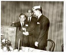 ["Black and white photograph of Carl Albert shaking hands with D.A.V Representative  Paul Freeman, along with an unidentified man in the background, at a Disabled American Veterans state convention dinner. 1970."]