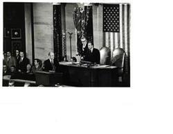 ["Black and white photograph of Carl Albert standing at the Speaker's chair. Gerald R. Ford is also present."]