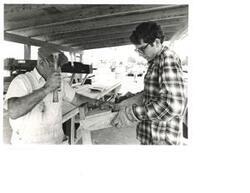 ["Black and white photograph of two unidentified men in makeshift outdoor work area/shelter."]