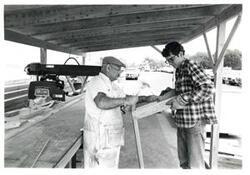 ["Black and white photograph of 2wo unidentified men in makeshift outdoor work area/shelter."]