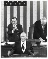 ["Black and white photograph of Gerald R. Ford standing at the speaker's podium. Carl Albert and James O. Eastland applauding behind him. Joint Session of Congress."]