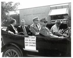 ["Black and white photograph of a car in a parade carrying Carl Albert, Henry Bellmon, Rear Admiral Christman, and Captain Chambliss in McAlester, Oklahoma on Armed Forces Day. 1974"]