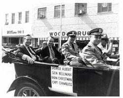 ["Black and white photograph of a car in a parade carrying Carl Albert, Henry Bellmon, Rear Admiral Christman, and Captain Chambliss in McAlester, Oklahoma on Armed Forces Day. 1974"]