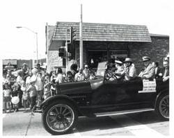 ["Black and white photograph of a car in a parade carrying Carl Albert, Henry Bellmon, Rear Admiral Christman, and Captain Chambliss in McAlester, Oklahoma on Armed Forces Day. 1974"]
