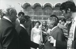 ["Black and white photograph print of Carl Albert shaking hands with a man. Mary Albert and others are in the background. This is during the House delegation trip to USSR, Romania, Yugoslavia. August 1975"]