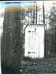 ["Black and white photograph of an outhouse in a wooded area."]