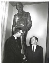 ["Black and white photograph of Carl Albert and an unidentified man, shaking hands, under a statue of Will Rogers."]