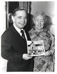 ["Black and white photograph of Carl Albert with Mrs. Lottie Ross holding a picture of an old schoolhouse with students in front. November 20, 1975.  Handwritten note on back of photograph, by Carl Albert, noting that the person in the photo was his first grade teacher, her age, and which child he is in the schoolhouse photograph in frame of picture."]
