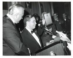 ["Black and white photograph of Carl Albert standing at a podium holding a recognition plaque."]